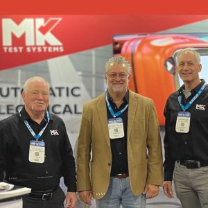 Three members of MK Test Systmes Americas team standing in front of a trade show booth, smiling at the camera.