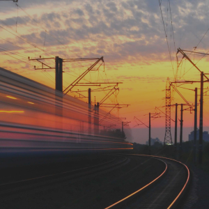 A train line is shown at sunset. A train rushes past in a blue. The sky is blue, orange, yellow, pink and purple. There are several overhead cables.