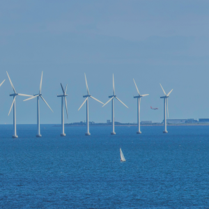 Offshore wind turbines positioned in a line. The water is calm and the sky is blue with no clouds.